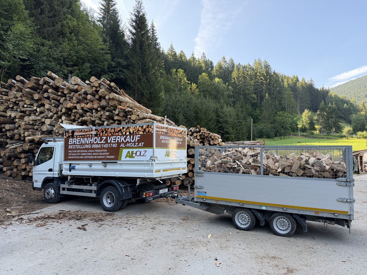 Weißer Klein-LKW mit beschrifteter Kipp-Pritsche voller Scheitholz, dahinter
ein zweiachsiger Anhänger mit Gitteraufsatz, ebenfalls beladen